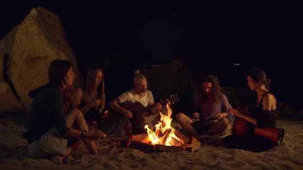 Friends Speaking Smiling Playing on Instruments Sitting Near Bonfire at Night Beach alt