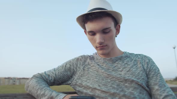 Young Attractive Guy in a Hat Reads Messages on His Phone and Smiles alt