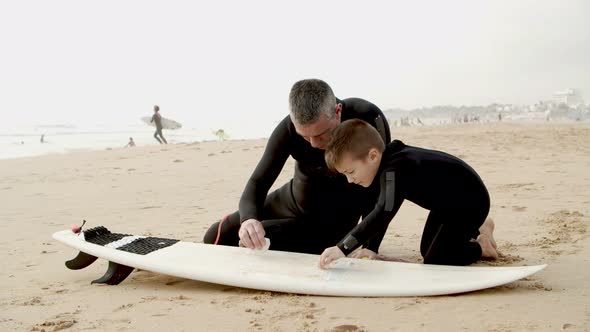 Father and Son Waxing Surfboard on Beach alt