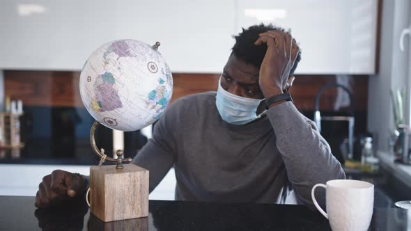 Worried African American Black Man Looking at Globe in His Home with Medical Mask. Traveling During alt
