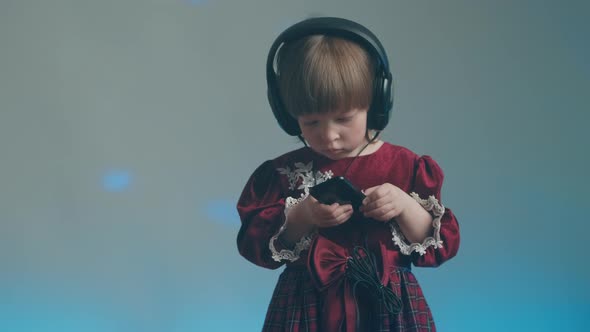 Little Girl in Big Headphones and Vintage Dress is Listening to Music on Phone