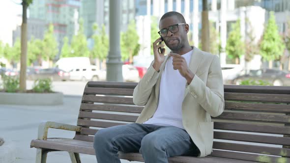 African Man Talking on Phone While Sitting Outdoor on Bench alt