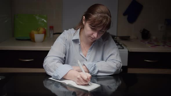 Woman Sits in the Kitchen at Home and Makes Plans Writes in a Notebook in the Evening