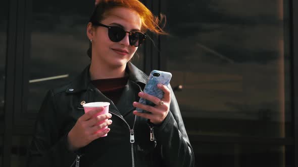 Young Woman Drinking Coffee on the Street, Typing Texting a Mobile Phone, While Walking in an Urban alt
