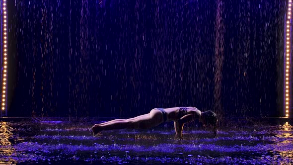 Beautiful Young Woman Stands in the Kumbhakasana Pose with Studio Blue Light and Water Rain. Female alt