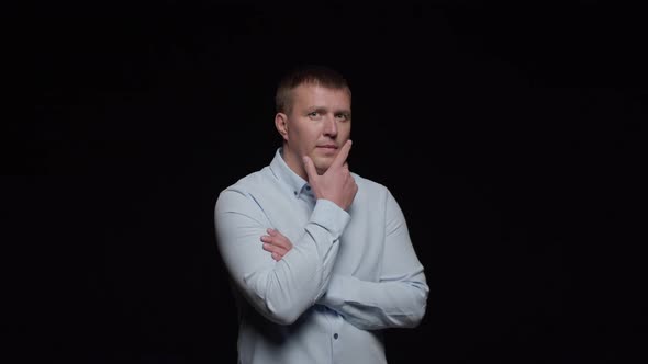 A young man in a white shirt poses on a black background, crosses his hands to squint his chin alt
