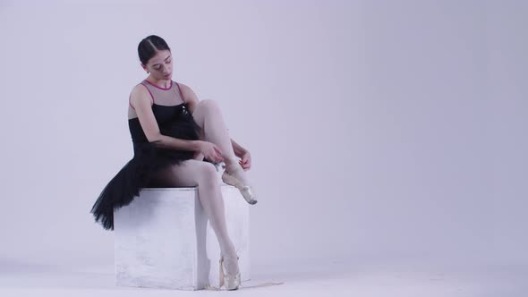 Young Woman Ballerina in Black Dress Putting on Pointe Shoes in the White Studio alt