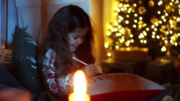 Cute Little Girl Is Lying on Bed and Write Letter To Santa in Room Decorated for Christmas alt