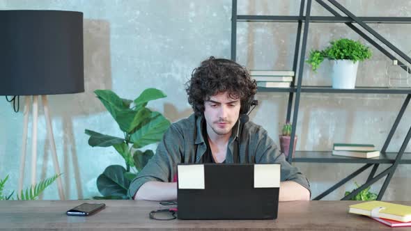 Young Curly Haired Man Using Laptop Computer with Headset to Talking Online alt