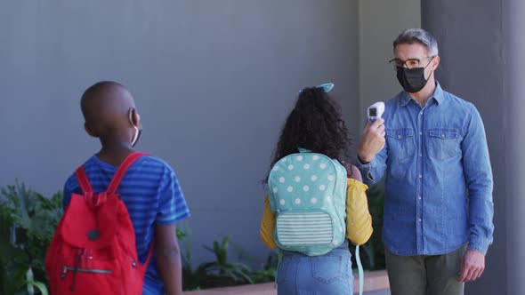 Diverse male teacher measuring temperature of schoolchildren, all wearing face masks alt