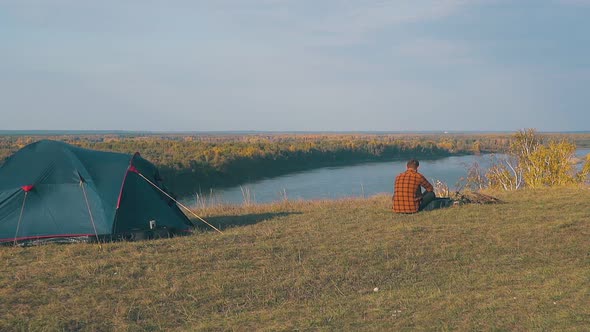 Guy in Orange Shirt Sits at Bonfire By Blue Tent in Morning alt