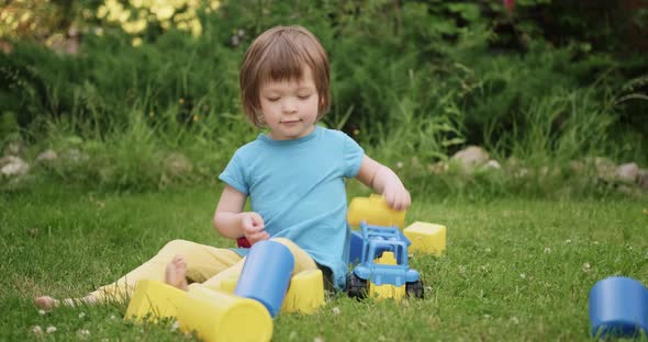 Small Cute Child is Playing with Toy Car and Cubes Sitting on Green Grass
