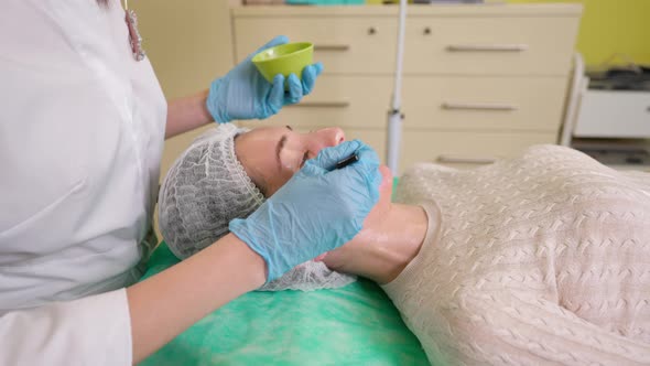 Beautician Applying Fruit Acid with Brush From Bowl on Face Skin of Young Woman alt