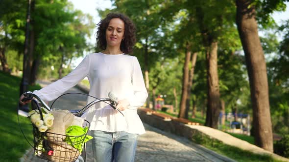 Young Woman in a White Tshirt and Blue Jeans Walking Holding Her City Bicycle's Handlebar with alt