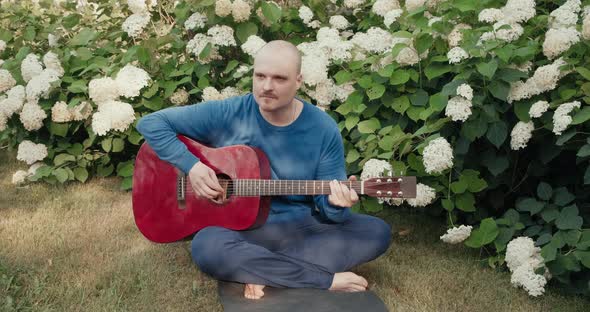 Caucasian Man is Sitting in a Park Among Flowers and Playing Red Acoustic Guitar