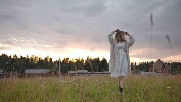 Gentle Woman in White Dress Walking in Rural Field Near Village alt