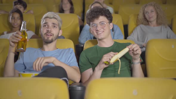 Unmannered Young Men Drinking Beer and Eating Banana in Movie Theatre. Portrait of Rude Guys alt