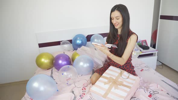 Happy Smiling Woman Holding Gift Card and Sitting on Bed with Balloons and Confetti alt