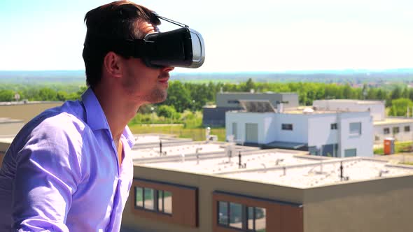 Young Man Uses Virtual Reality Glasses on the Balcony - Buildings and Nature in the Background alt