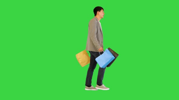 Young Asian Man Walks with Colored Packages and Smiles on a Green Screen Chroma Key alt