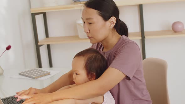 Asian Woman Sitting with Baby Daughter and Working on Laptop alt