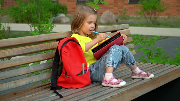 Girl Sits on Bench with Backpack in Schoolyard and Does Homework