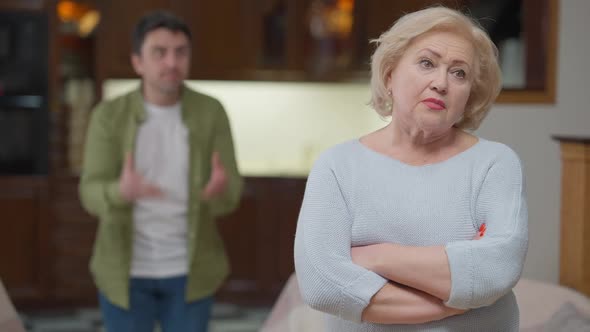 Portrait of Sad Disappointed Senior Woman Standing with Crossed Hands As Nervous Angry Man Yelling alt