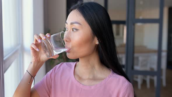 Attractive Asian Woman is Drinking Water From Glass Sitting on Sofa at Home Restores Water Balance alt