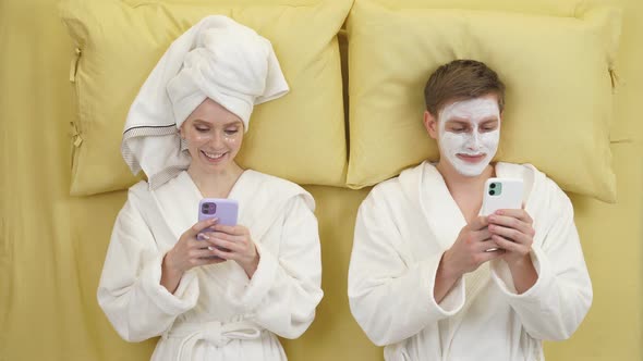 Relaxed Man And Woman After Beauty Procedures At Home Lying On Bed Having Rest Using Mobile Phones