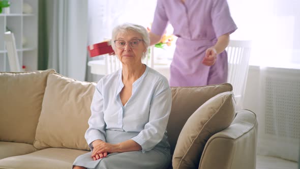 Long haired blonde in purple dress gives small box with present to aged lady alt