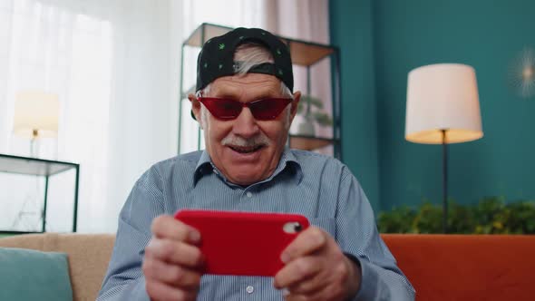 Worried Funny Senior Old Grandfather Man Playing Shooter Online Video Games on Mobile Phone at Home alt