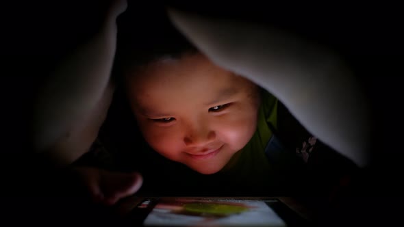 Night shot of Happy little asian boy playing with tablet or smartphone alt