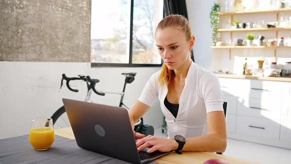 Young Woman is Typing on Laptop Sitting at Table in Kitchen at Home alt