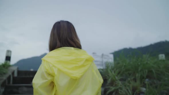 Young Asian woman feeling happy playing rain while wearing raincoat standing near lake. alt