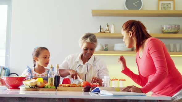 Mother looking recipe book while grandmother and granddaughter chopping vegetables alt