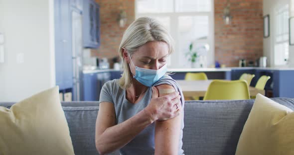 Portrait of happy senior caucasian woman wearing face mask in living room with bandage on her arm alt