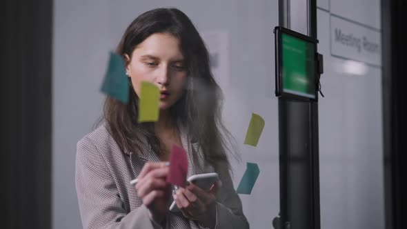 Beautiful Concentrated Woman Checking Schedule in Smartphone Writing with Pen on Sticky Notes alt