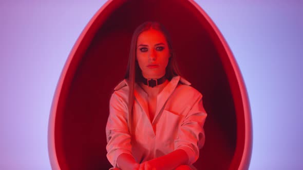Elegant Woman Posing in Egg Chair in Studio with Neon Light alt