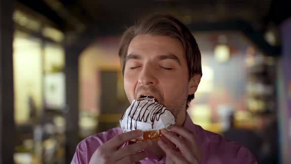 Closeup of a Handsome Man Greedily Biting a Cake with Icing and Closes His Eyes with Pleasure