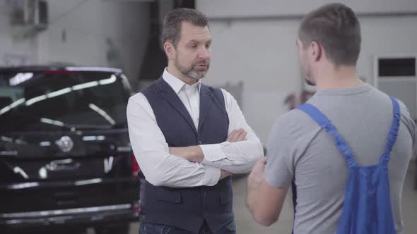 Portrait of Unsatisfied Adult Caucasian Man Talking with Auto Mechanic in Repair Shop. Handsome alt
