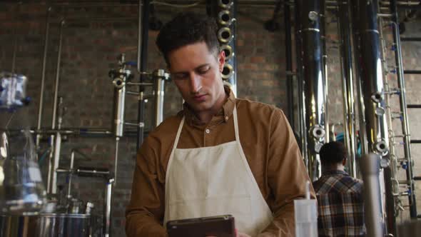 Caucasian man working at gin distillery, using digital tablet, wearing apron alt
