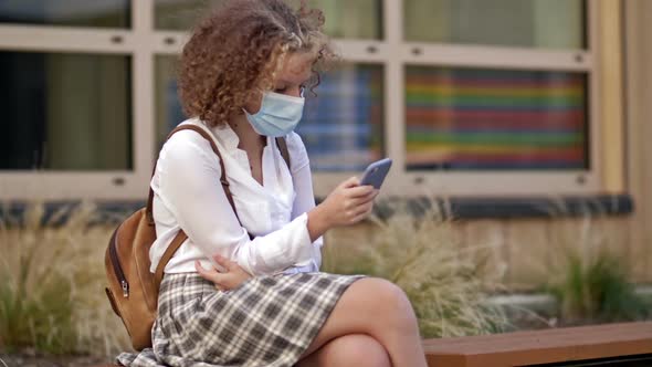 Teenage Schoolgirl in Protective Mask Sits on a Bench Near the School Building with a Smartphone in alt