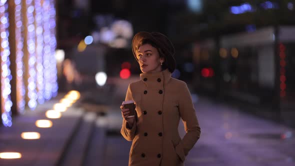 Beautiful Woman in Hat and Coat Drinks Coffee and Walks in the Night City