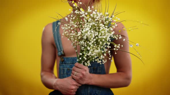 Unrecognizable Young Queer Man in Denim Overalls Posing with White Flowers at Yellow Background alt