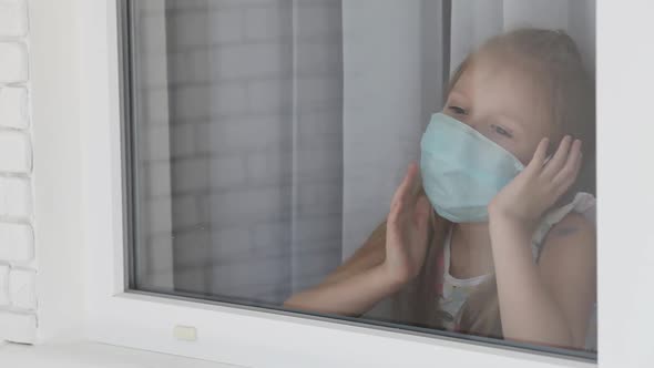 Little girl in a medical mask sad look behind a window in a house. Quarantine Stay home