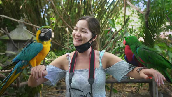 Asian Woman Tourist with Photo Camera and Medical Mask Holds Two Parrots on Both Hands