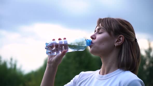 Young Beautiful Athletic Girl in a White T-shirt Is Very Tired and Drinks Water From a Transparent alt