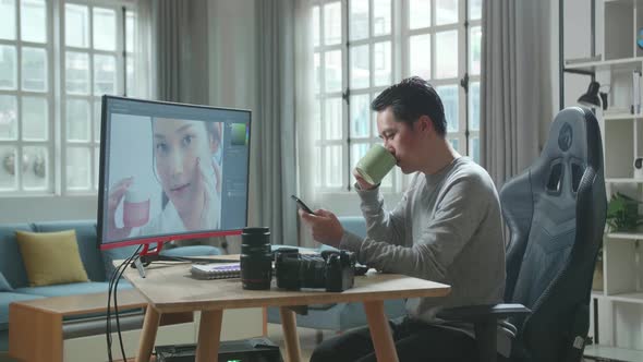 Asian Cameraman Using Mobile Phone And Drink Coffee While Using Computer For Editing Photo At Home alt