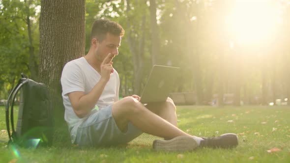 Young Student on the Grass in the Park Sits Under a Tree and Cheerfully Works with a Laptop alt