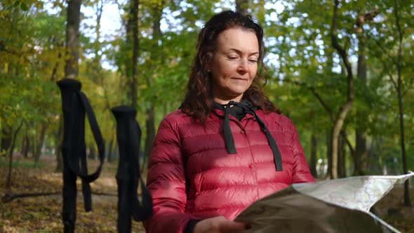 Portrait of Thoughtful Caucasian Mature Woman Examining Paper Map Standing in Autumn Park with Poles alt